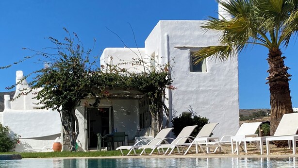 A poolside area with white buildings, a palm tree, and lounge chairs. - Paros, Drios Golden Beach: beautiful villa, large private pool, unique sea view (chrisi akti)