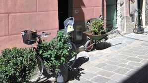 Una calle estrecha con un edificio rosa, una bicicleta, un carro de mano y una planta en maceta.