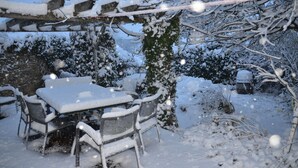 A snow-covered outdoor seating area with a table and chairs, surrounded by snow-laden trees and branches. - COTSWOLD COTTAGE IN A QUIET VILLAGE WITH LARGE GARDEN (Great Rissington)