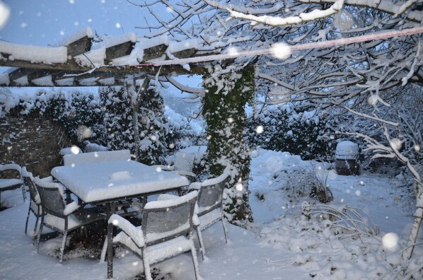 Un área de estar al aire libre cubierta de nieve, con una mesa y sillas, rodeada de árboles y ramas cargadas de nieve.