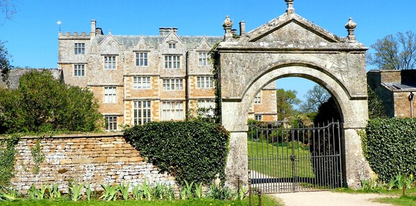 A historic stone building with a large arched entrance, a brick wall with ivy, and a well-maintained lawn. - COTSWOLD COTTAGE IN A QUIET VILLAGE WITH LARGE GARDEN (Great Rissington)