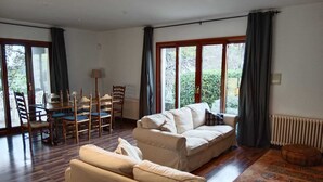 Uma sala de estar ampla com um sofá bege, uma mesa de jantar com cadeiras e grandes janelas com cortinas.