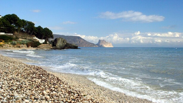 Platja de petits pedres amb aigua blava cristal·lina i muntanyes a la distància.