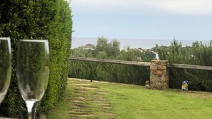 A table with a charcuterie board and two glasses of wine, overlooking a grassy area with a stone wall and trees.