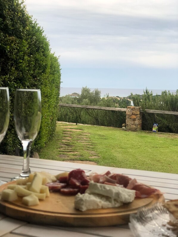 Una mesa con una tabla de embutidos y dos copas de vino, con vista a una zona verde con un muro de piedra y árboles.