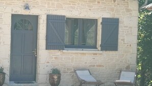 A stone building with a closed door, two windows with shutters, and two lounge chairs outside.