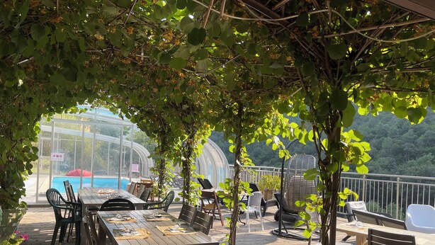 An outdoor patio with wooden tables and chairs, surrounded by greenery and a view of a swimming pool and forested hills.