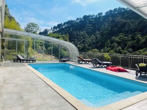 Une piscine dotée de chaises longues et d’un patio vitré, le tout entouré d’une végétation luxuriante et de collines.