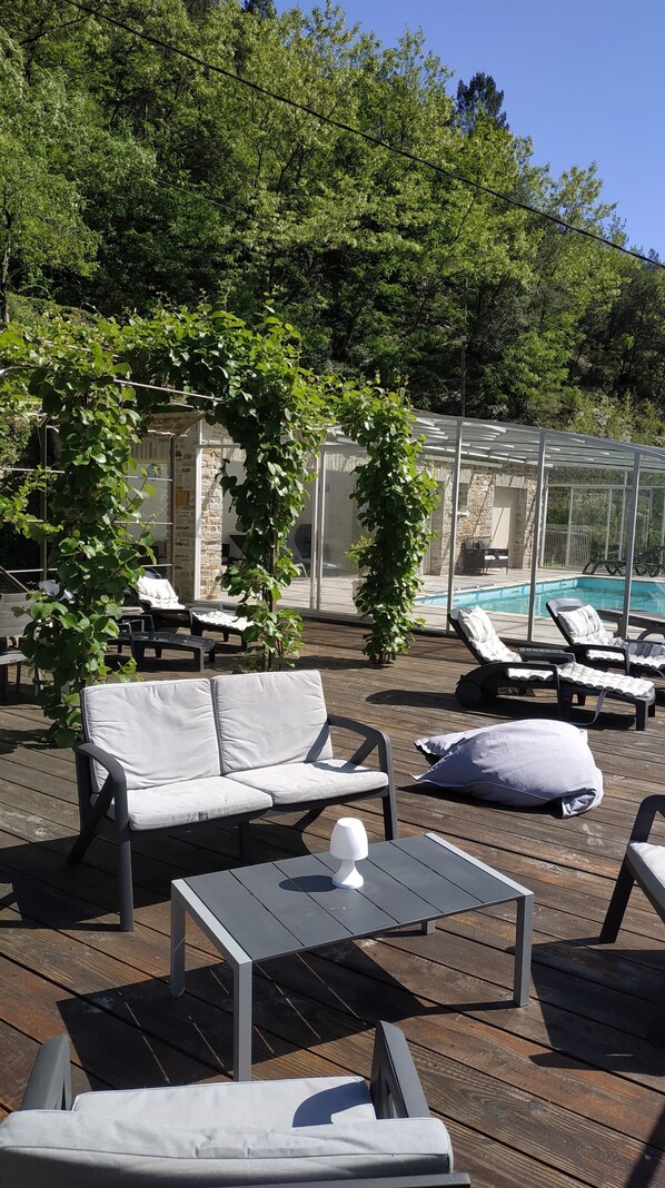 Une terrasse en bois avec des sièges extérieurs, une piscine à moitié couverte par une paroi de verre et une végétation luxuriante.