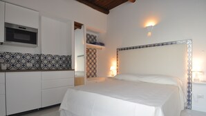Un dormitorio con cama, mesita de noche, televisor montado en la pared y una zona de cocina con armarios y microondas.