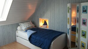 A bedroom with a sloped ceiling, a bed with a blue blanket, a nightstand with a lamp, and a wardrobe with pictures. - Penmarch: Country Cottage / Gite - Penmarc'h (Penmarch)
