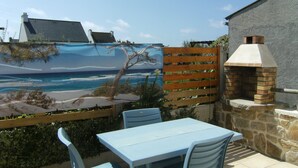 A patio with a table and chairs, a brick oven, and a wooden fence with a beach mural. - Penmarch: Country Cottage / Gite - Penmarc'h (Penmarch)