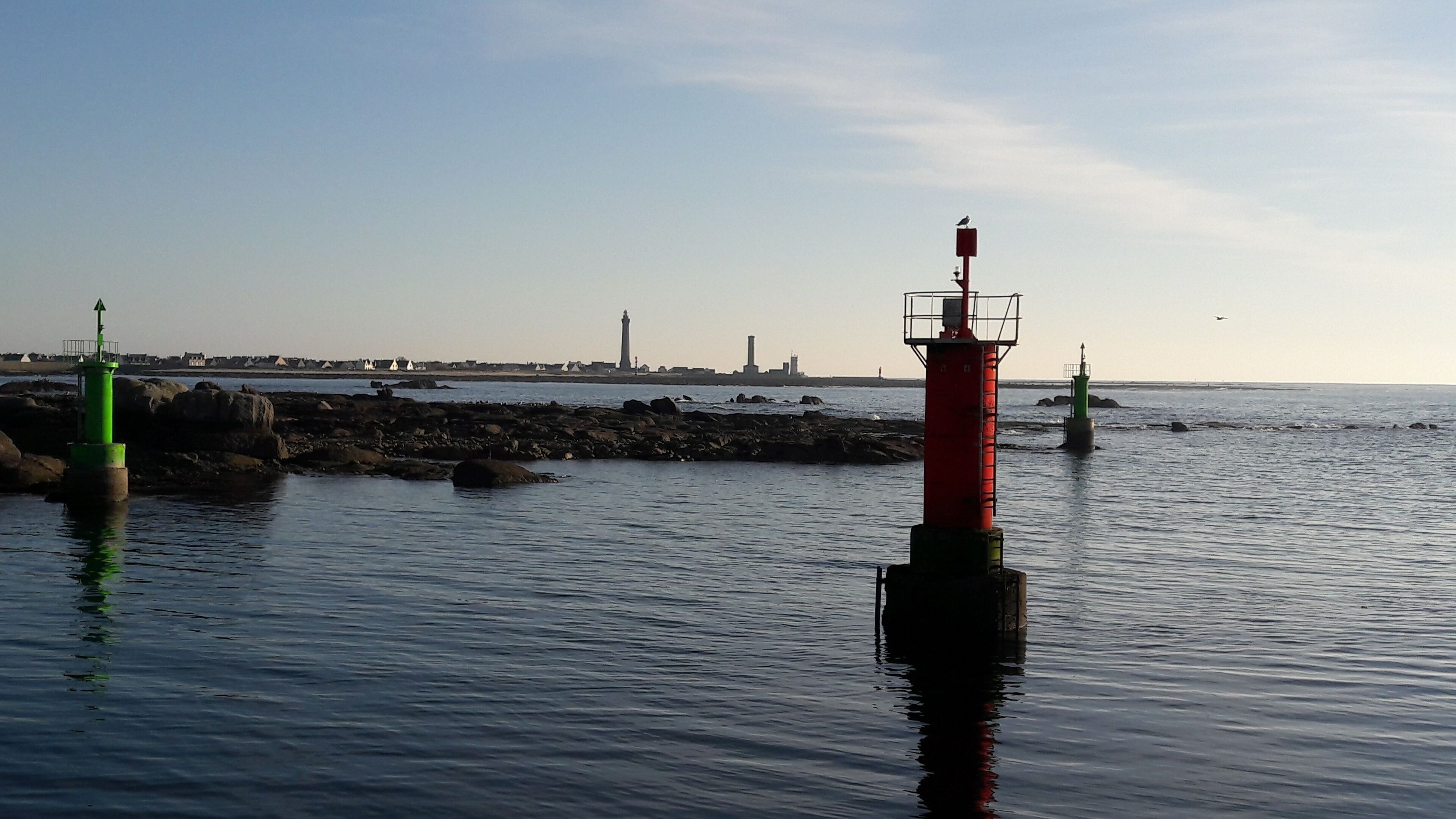Un paysage côtier avec un phare rouge bien visible, une bouée verte et une côte rocheuse.