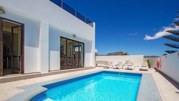 Un área de piscina moderna con una piscina azul cristalina, paredes blancas y vistas a las montañas.