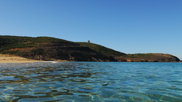 A coastal landscape with clear water, a sandy beach, and a hill with a structure on top. - TORRE DEI CORSARI, THE PEARL OF THE GREEN COAST - IUN Code: P7053 (Arbus)