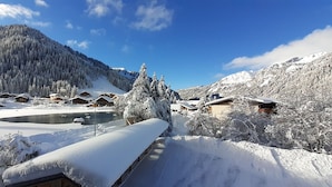 A snow-covered mountain village with wooden chalets, a pond, and a clear blue sky.