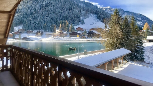 A view from a wooden balcony overlooking a lake with snow-covered houses and mountains in the background.