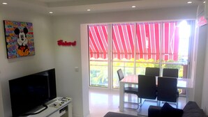 A modern living room with a flat-screen TV, a dining area with a table and chairs, and a balcony with red curtains. - Cannes - Sea front apartment, beach front, pool, garden, internet, aircon (Cannes)