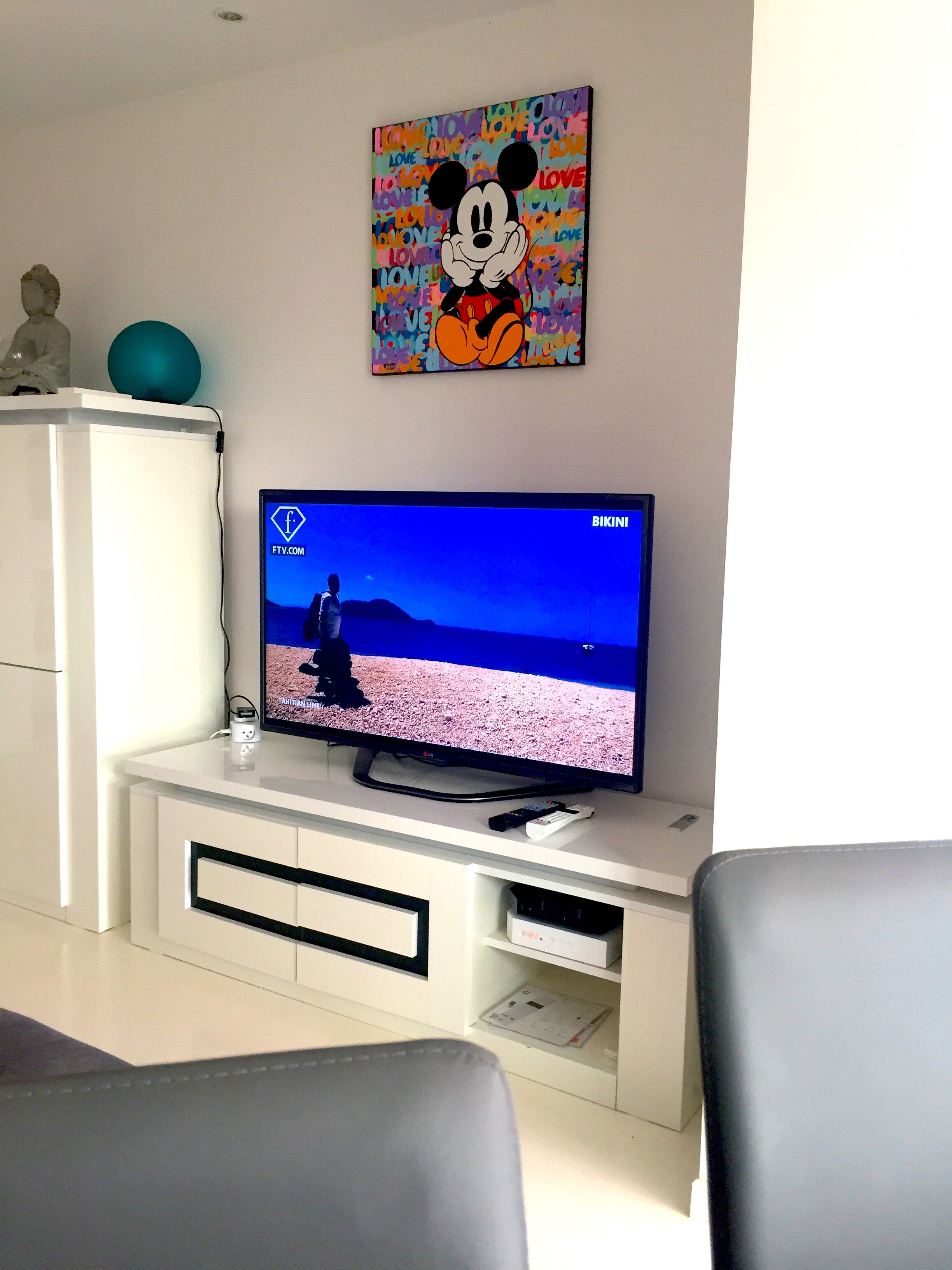 Un téléviseur posé sur un meuble blanc, accompagné d’une télécommande, d’une sphère décorative et d’un tableau accroché au mur représentant un personnage de dessin animé.