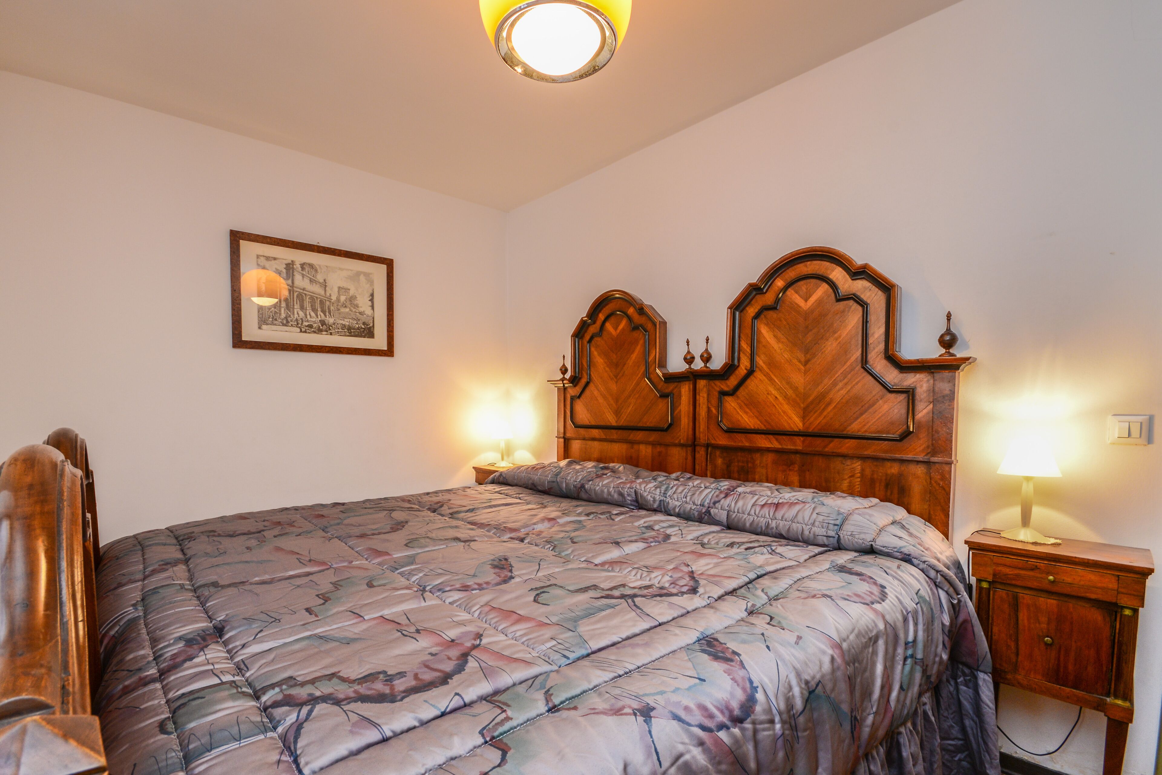 A neatly made bed with a floral-patterned bedspread, a wooden headboard with decorative carvings, and two bedside lamps.