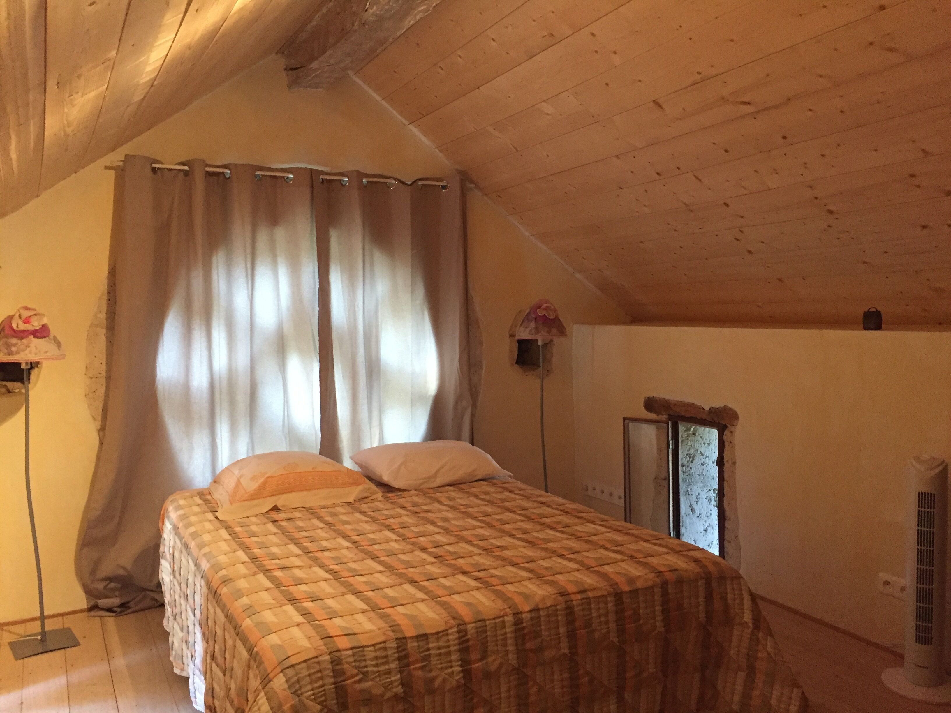 A bedroom with a wooden ceiling, a bed with a plaid bedspread, a window with curtains, and a wall-mounted heater.