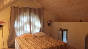 A bedroom with a wooden ceiling, a bed with a plaid bedspread, a window with curtains, and a wall-mounted heater.