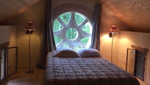 A bedroom with a large circular window, a bed with white pillows, and two floor lamps. - Charming property with its chapel and towers (Lannes)