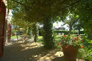 Eine Terrasse mit Tisch und Stühlen, umgeben von Grünpflanzen und einem großen Topf mit roten Blüten.