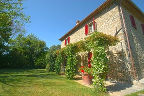 Ein Steinhaus mit roten Fensterläden, ein Garten mit Gras und blühenden Pflanzen, und ein klarer, blauer Himmel.
