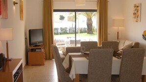 A dining area with a table and chairs, a television on a stand, and a view of the outdoors through a glass door. - Luxury 2 Bed Apartment with pool near to beach (Porto De Mos)