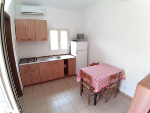 Cozinha compacta com armários de madeira, fogão a gás, pia e um frigorífico pequeno. Há uma janela com persianas, um micro-ondas em cima do frigorífico e uma mesa com toalha xadrez.