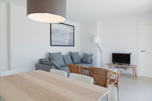 A modern living room with a wooden dining table, grey sofa, wicker chairs, a flat-screen TV, and a framed picture on the wall. - Penthouse Sea Views · AirCon · Pool · Parking · UHC ACACIAS 263 (Salou)
