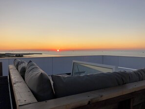 A balcony with a view of the sunset over the sea, a cushioned seating area, and a wooden railing.