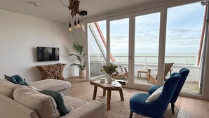 Sala de estar com sofá, cadeiras, mesa de centro e televisão. Há uma janela grande com vista para o oceano.