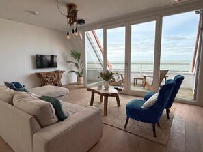 A living room with a sofa, chairs, a coffee table, and a TV. There is a large window with a view of the ocean.
