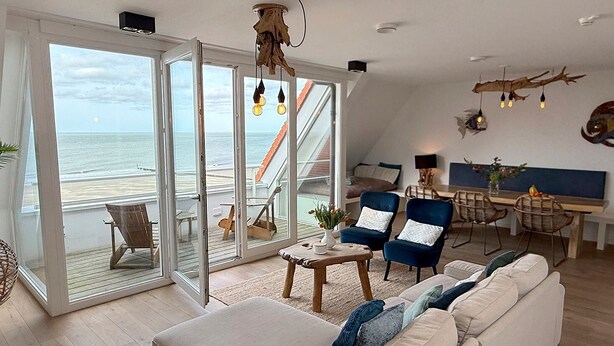 Uma sala de estar espaçosa com uma grande porta de vidro deslizante que dá para uma varanda com vista para o mar, um espaço de estar aconchegante com sofá e poltronas, e uma mesa de jantar com cadeiras.