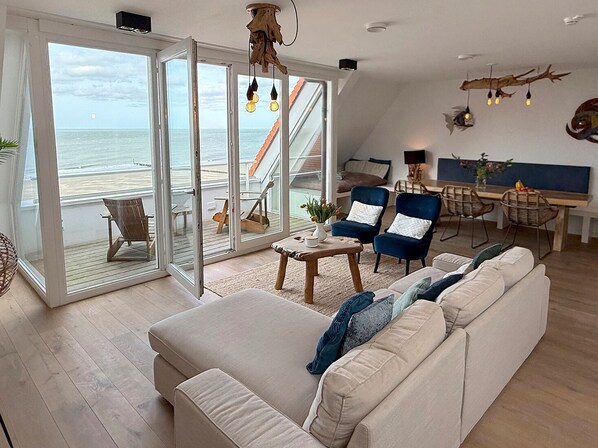 A spacious living area with a large sliding glass door leading to a balcony overlooking the sea, a cozy seating area with a sofa and chairs, and a dining table with chairs.