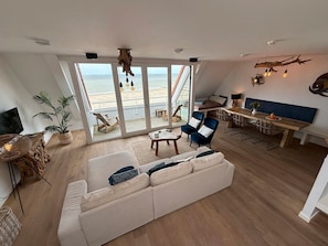 Amplia sala de estar con un sofá grande, una mesa de comedor y sillas, un balcón con vista a la playa y una zona de cocina.