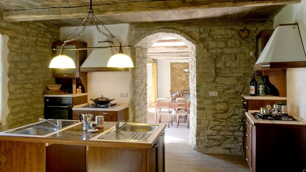 Una cocina rústica con paredes de piedra, armarios de madera y una isla central con lavamanos. Hay una estufa, una lámpara colgante y vista al área de comedor a través de un arco.