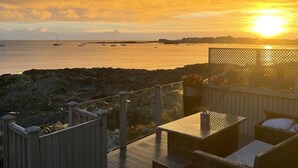 A deck with outdoor furniture overlooking a rocky coastline and the sea at sunset.