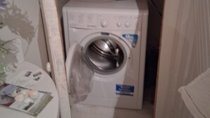 A compact laundry room with a front-loading washing machine, a small table with a magazine, and a wall-mounted shelf.
