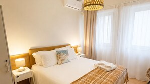 A hotel room with a bed, bedside tables, a lamp, an air conditioner, and a window with curtains. - Charming house in downtown Olhão (Olhão)