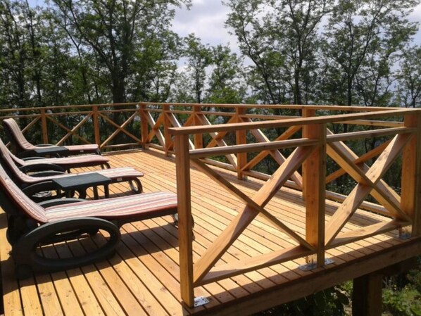 A wooden deck with lounge chairs and a table, surrounded by a railing, overlooking a forested area.