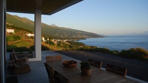 A wooden table and chairs on a terrace overlooking a coastal landscape with mountains in the background. -  AtlanticWindow - Modern House, Stunning View (São Roque do Pico)