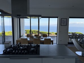 A modern kitchen with a gas stove, dining area with chairs, and large windows offering a view of the sea and mountains. -  AtlanticWindow - Modern House, Stunning View (São Roque do Pico)