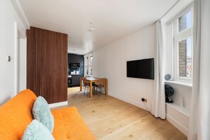 A modern living room with a wooden floor, a dining area with a table and chairs, a kitchen in the background, and a TV mounted on the wall. - PRIVATE APARTMENT - HEART OF SHOREDITCH (London)