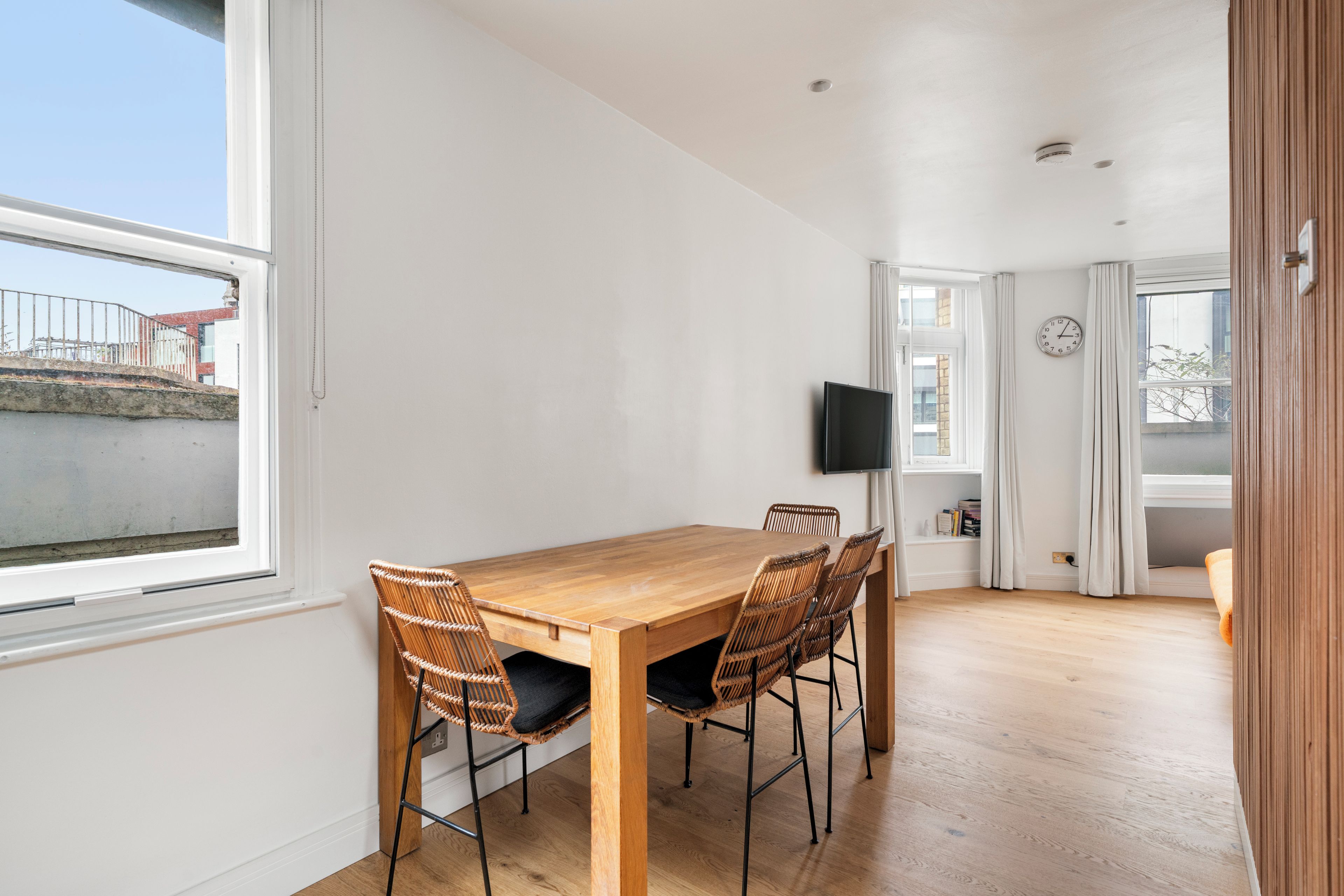 A modern dining area with a wooden table and wicker chairs, a flat-screen TV mounted on the wall, and a window with curtains.
