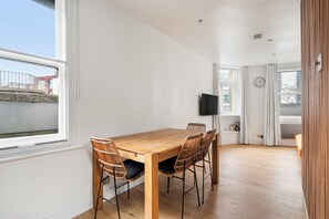 A modern dining area with a wooden table and wicker chairs, a flat-screen TV mounted on the wall, and a window with curtains. - PRIVATE APARTMENT - HEART OF SHOREDITCH (London)