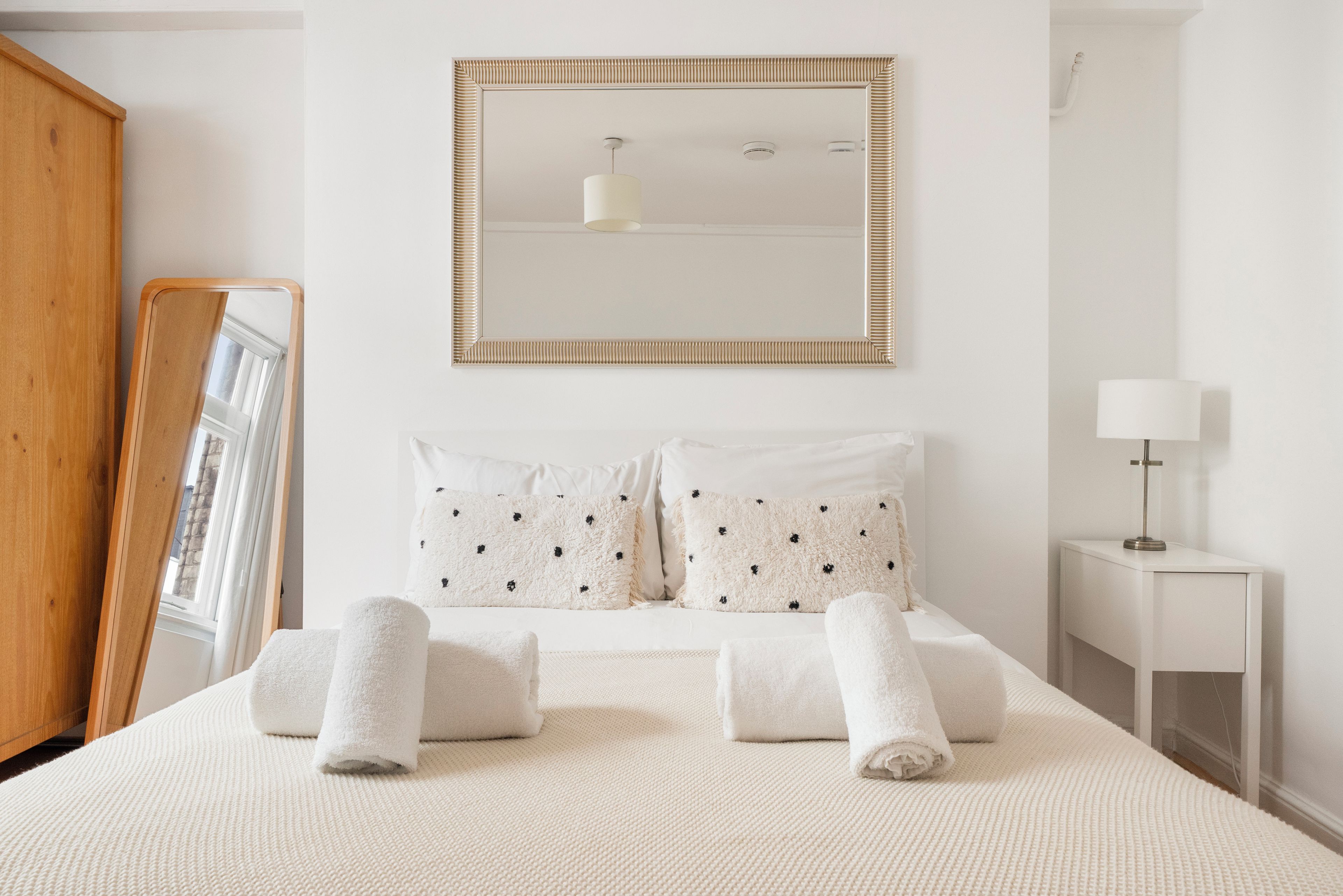 A neatly made bed with white linens and two pillows, a bedside table with a lamp, and a large mirror on the wall.