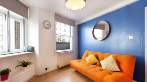 A room with a blue wall, a round mirror, a window with a view of buildings, a radiator, and a wooden floor. - PRIVATE APARTMENT - HEART OF SHOREDITCH (London)